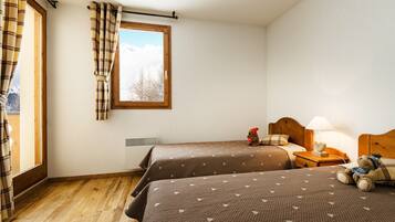 Une chambre avec deux lits, une fenêtre avec des rideaux, une table de chevet avec une lampe et un ours en peluche sur le lit de droite.