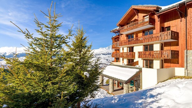 A wooden chalet with balconies, a stone entrance, and a snowy mountain backdrop.