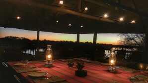 Un espace de restauration en plein air avec une nappe rouge, une table dressée pour un repas agrémenté de bougies et offrant une vue sur le coucher de soleil au-dessus d’une étendue d’eau.