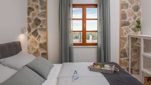 A bedroom with a stone wall, a bed with grey bedding, a window with curtains, and a bedside table with a tray of items. - Beautiful 3-BR villa with pool and sea view just above Rogoznica (Rogoznica)