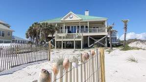 A beachfront house with a green roof, a balcony, and a sandy beach in the foreground.