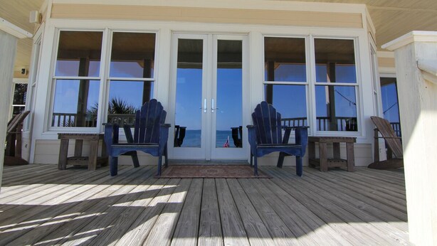 A wooden deck with two Adirondack chairs facing a large glass door leading to a balcony with a view of the ocean.