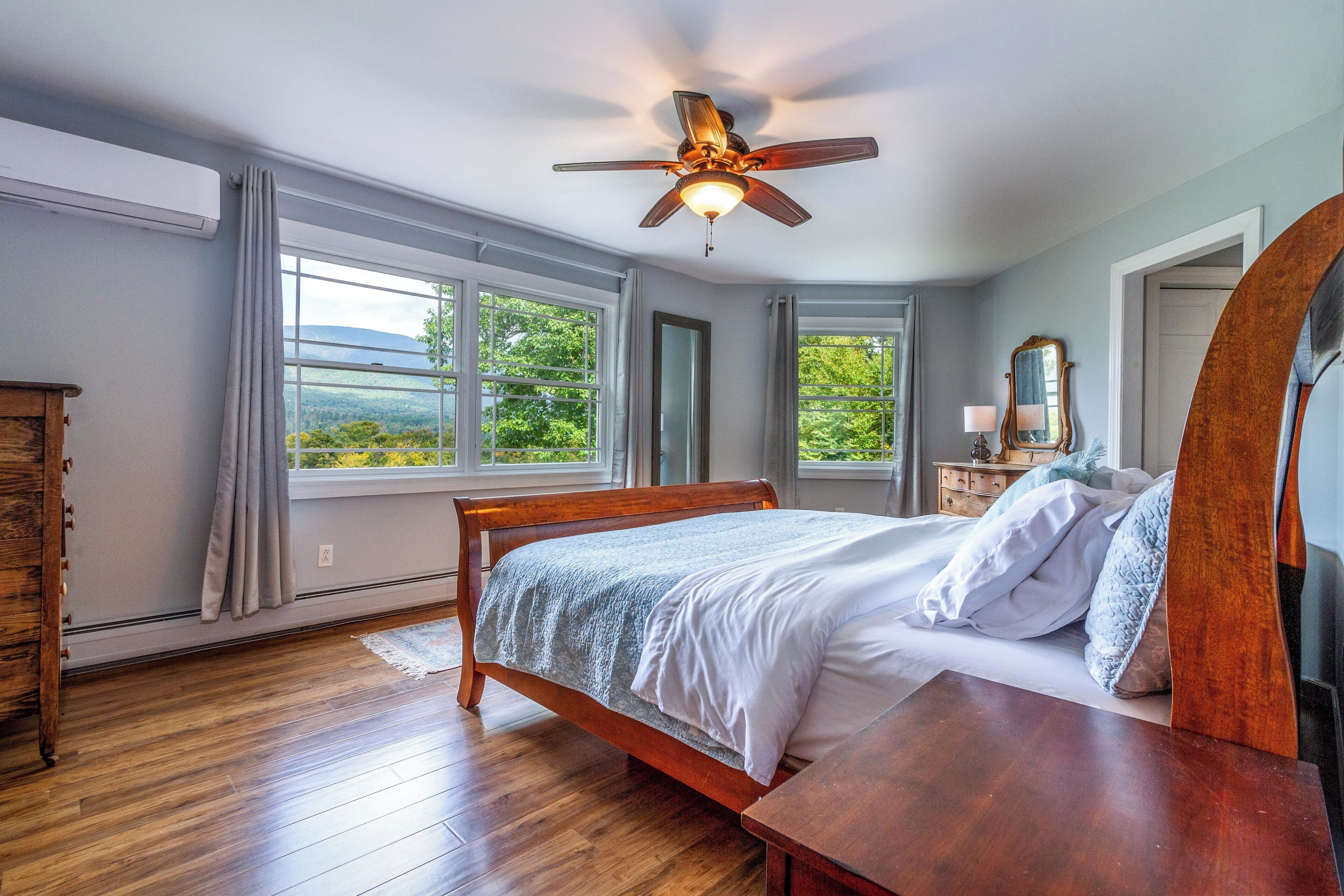 A bedroom with a wooden bed, a ceiling fan, large windows, and a view of greenery outside.