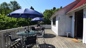 Une terrasse en bois avec une table et des chaises en verre, deux parasols bleus et un bâtiment blanc avec un toit rouge.