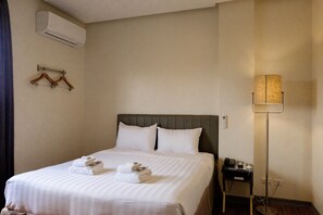 Una cama bien hecha con sábanas y toallas blancas, una mesita de noche con lámpara y un aire acondicionado instalado en la pared.