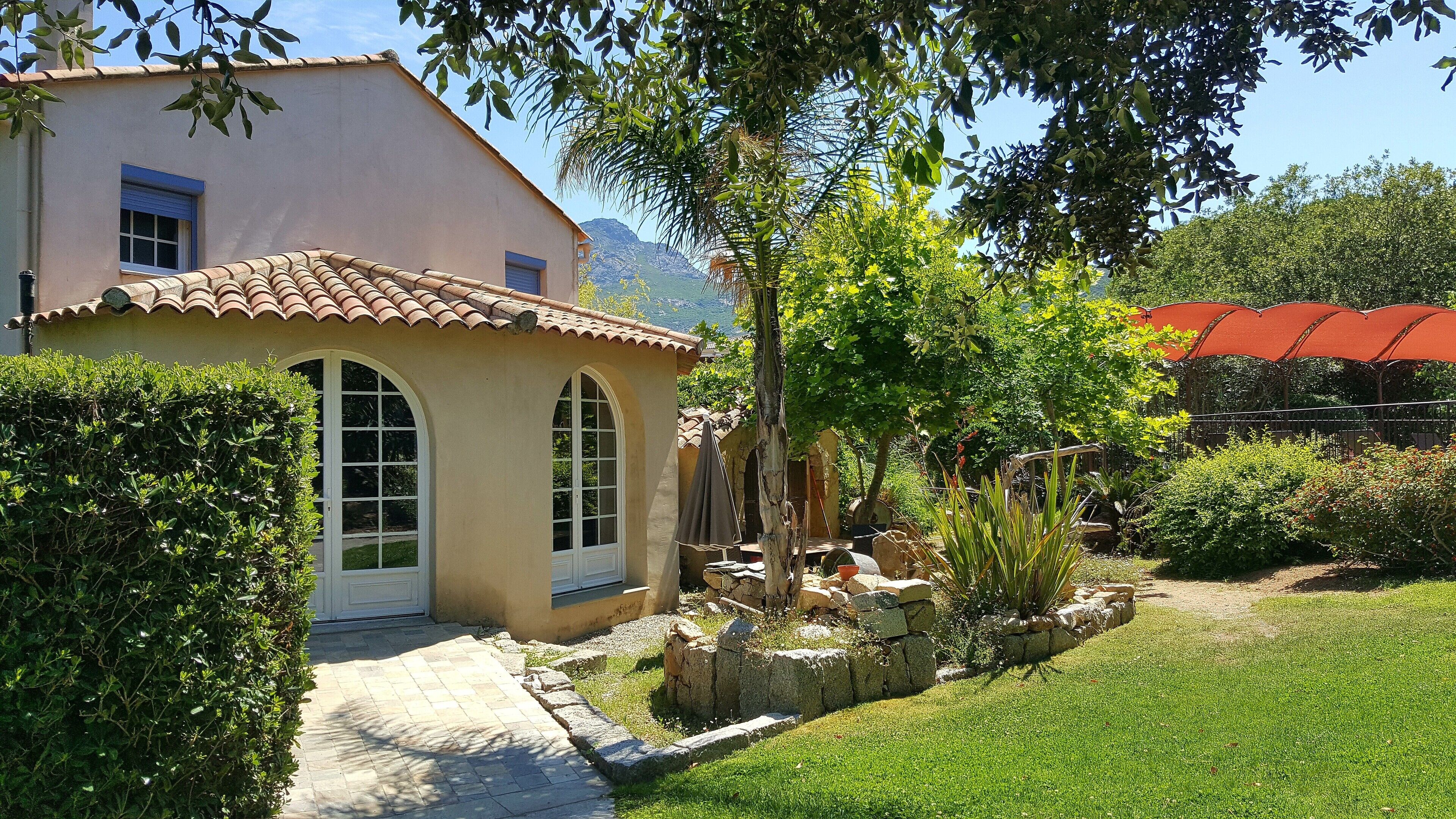 A house with a tiled roof, a small garden with a pathway, and a mountain in the background.