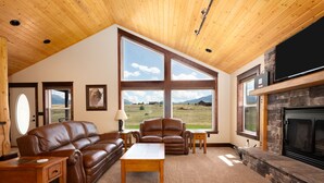 A spacious living room with a large window offering a view of a mountainous landscape, a leather sofa, a wooden coffee table, and a stone fireplace. - Yellowstone Trail - Great Home Overlooking the Yellowstone River (Pray)