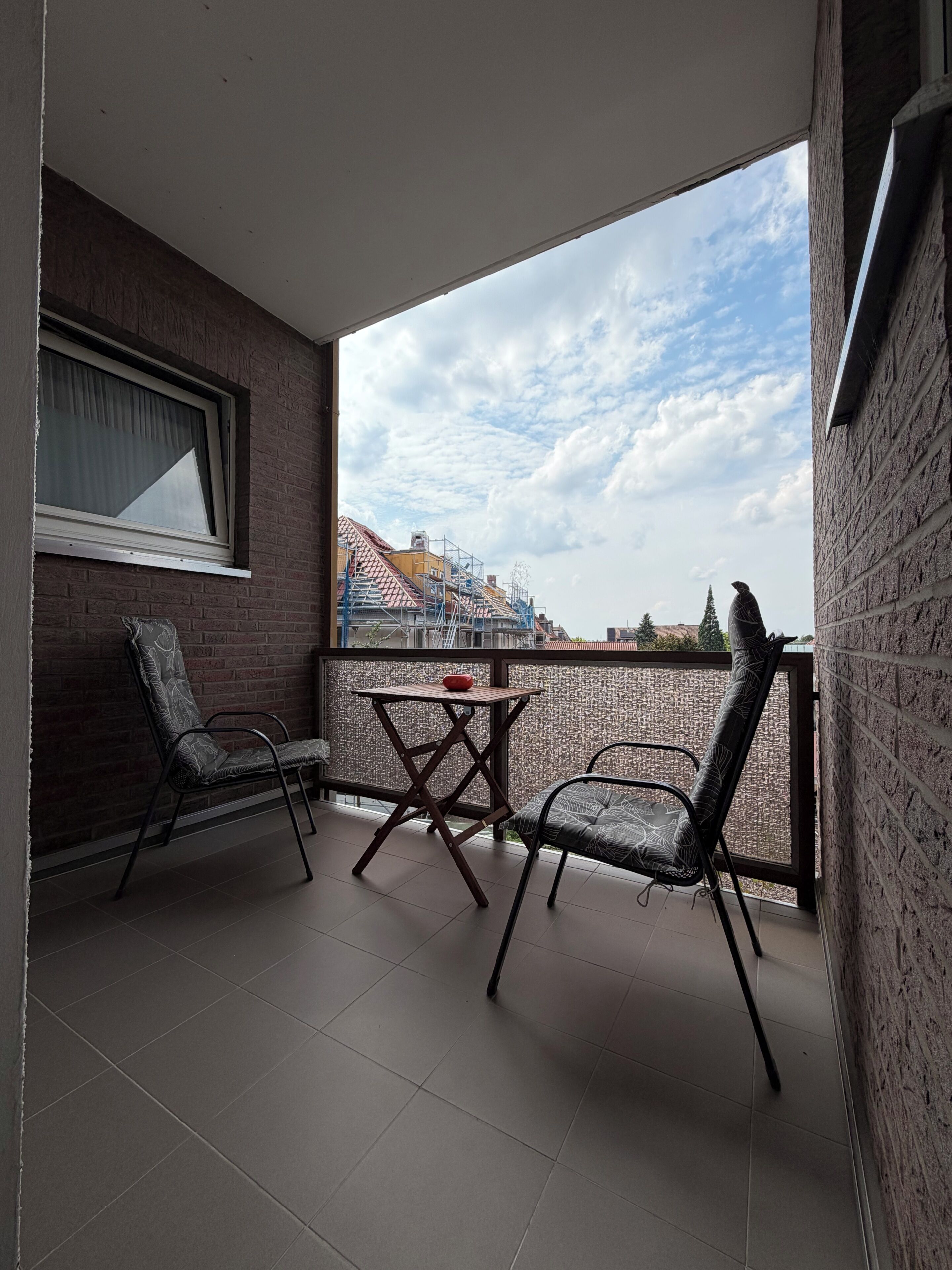 A balcony with two chairs and a table, a window with curtains, and a view of buildings and trees.