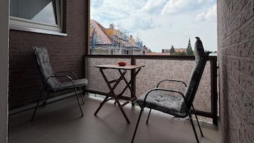 A balcony with two chairs and a table, a window with curtains, and a view of buildings and trees.