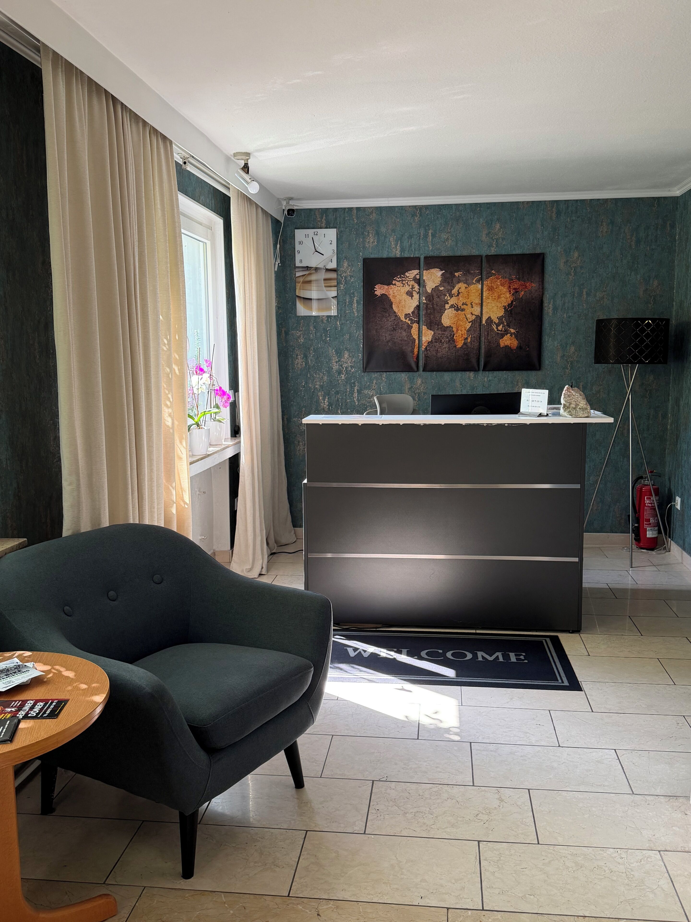 Ein modernes Wohnzimmer mit einem dunklen Sofa, einem Empfangstresen, einer Weltkarte als Kunstwerk und einer Uhr an der Wand.