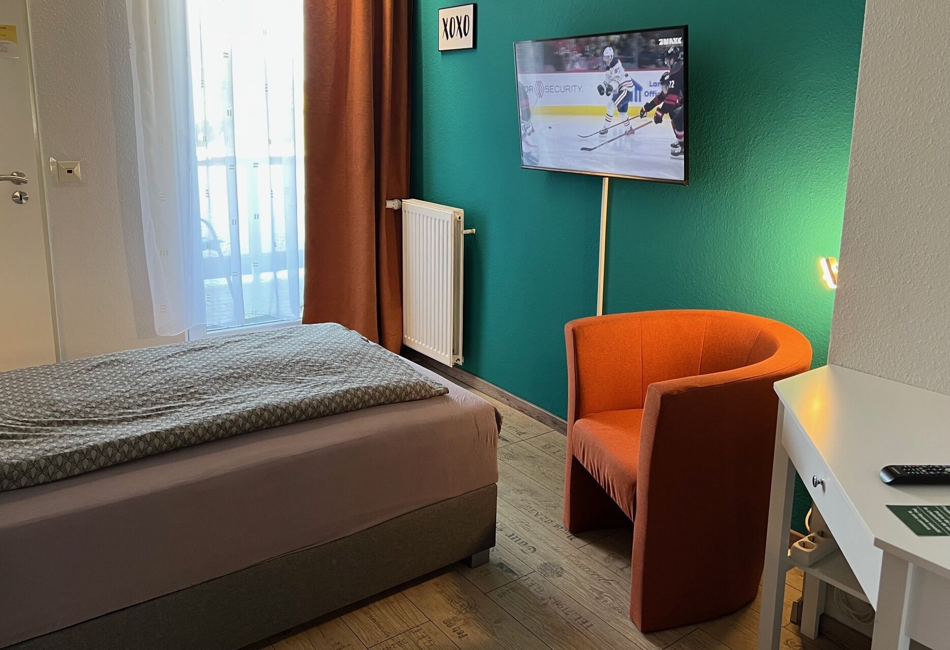 A hotel room with a bed, an orange armchair, a desk with a computer, a TV on the wall, and a window with curtains.