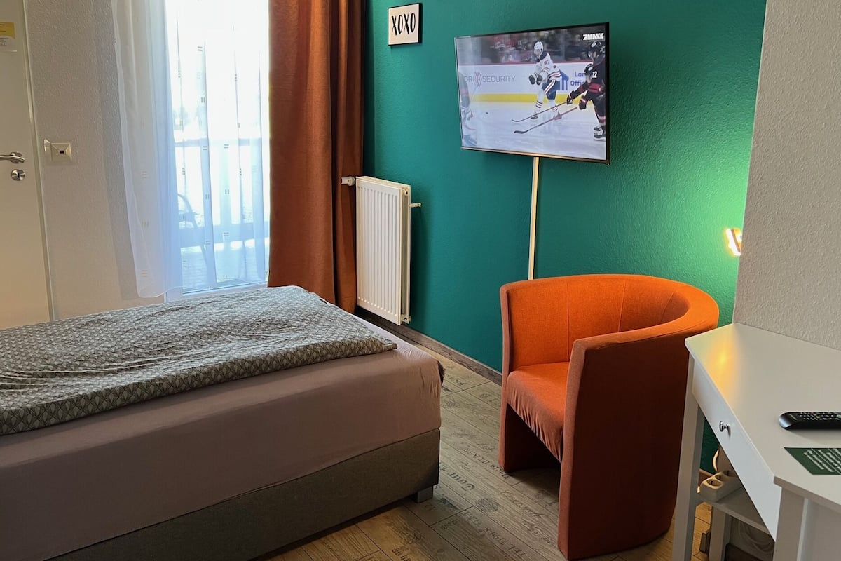 A hotel room with a bed, an orange armchair, a desk with a computer, a TV on the wall, and a window with curtains.