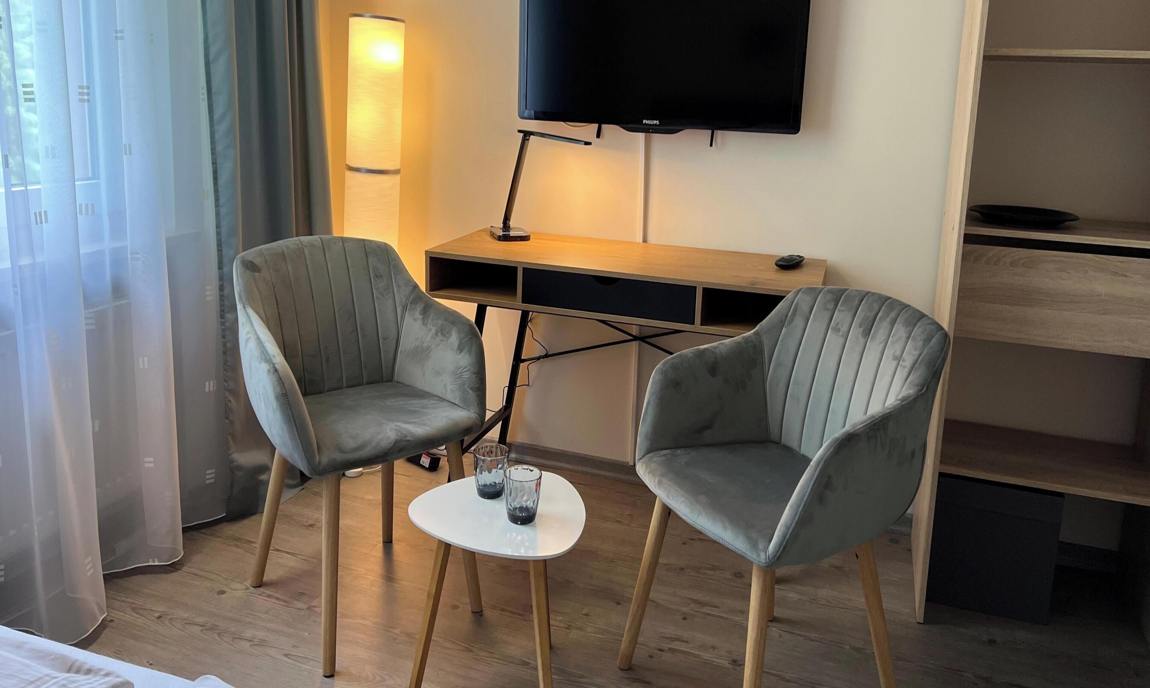 A room with two grey upholstered chairs, a small white table, a flat-screen TV, and a wooden desk.