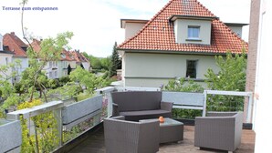 Eine Terrasse im Freien mit Korbmöbeln, einem Geländer und Blick auf benachbarte Häuser.