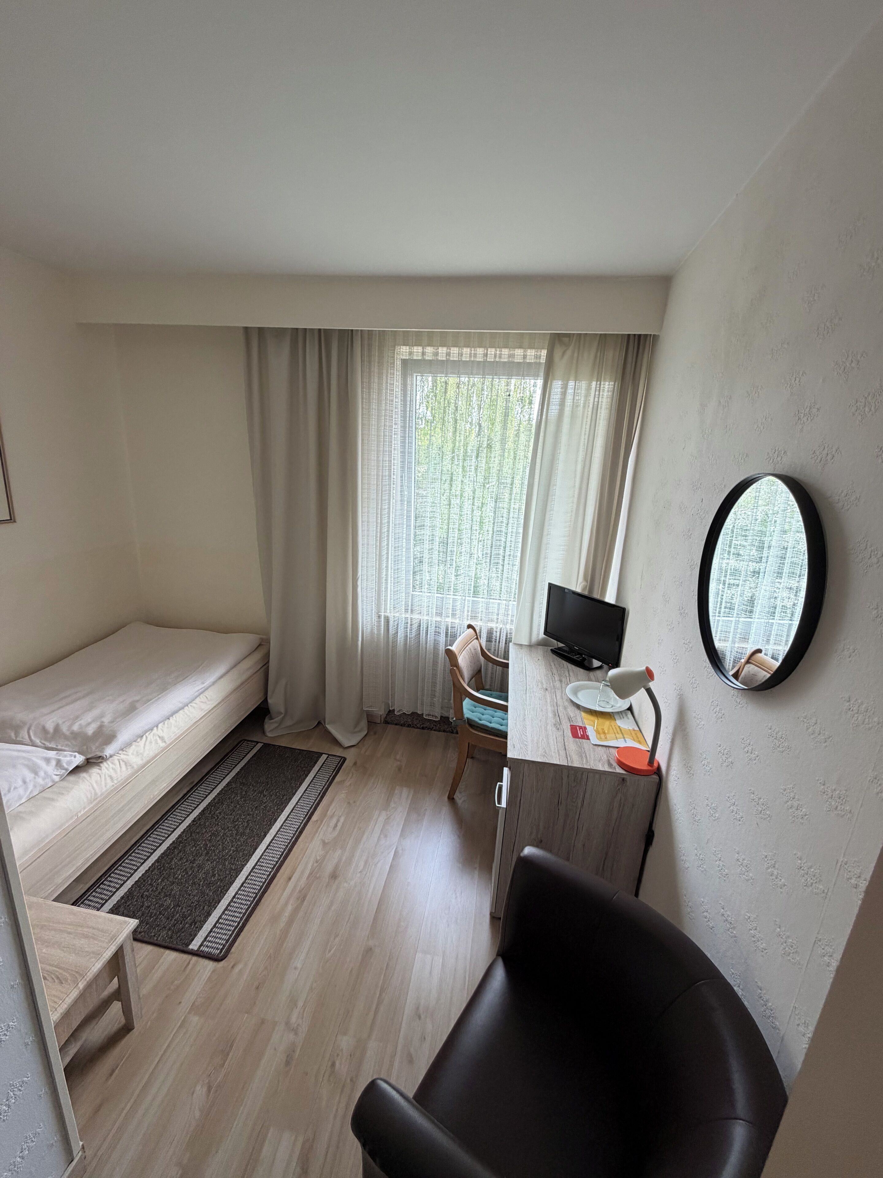 Et lille værelse med en enkelt seng, et skrivebord med computer og lampe, en stol og et vindue med gardiner.