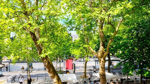 Ein Straßencafé mit Tischen und Stühlen unter Bäumen, ein rotes Gebäude im Hintergrund und eine Straße mit Fußgängern und Fahrrädern.