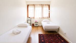 Ein kleines, sauberes Zimmer mit zwei Einzelbetten, einem Fenster mit durchscheinenden Vorhängen, einem kleinen Nachttisch und einem gemusterten Teppich.