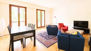 Ein geräumiger Wohnbereich mit Esstisch, Stühlen, einer Couch, einem roten Sessel, einem Fernseher und einem gemusterten Teppich.