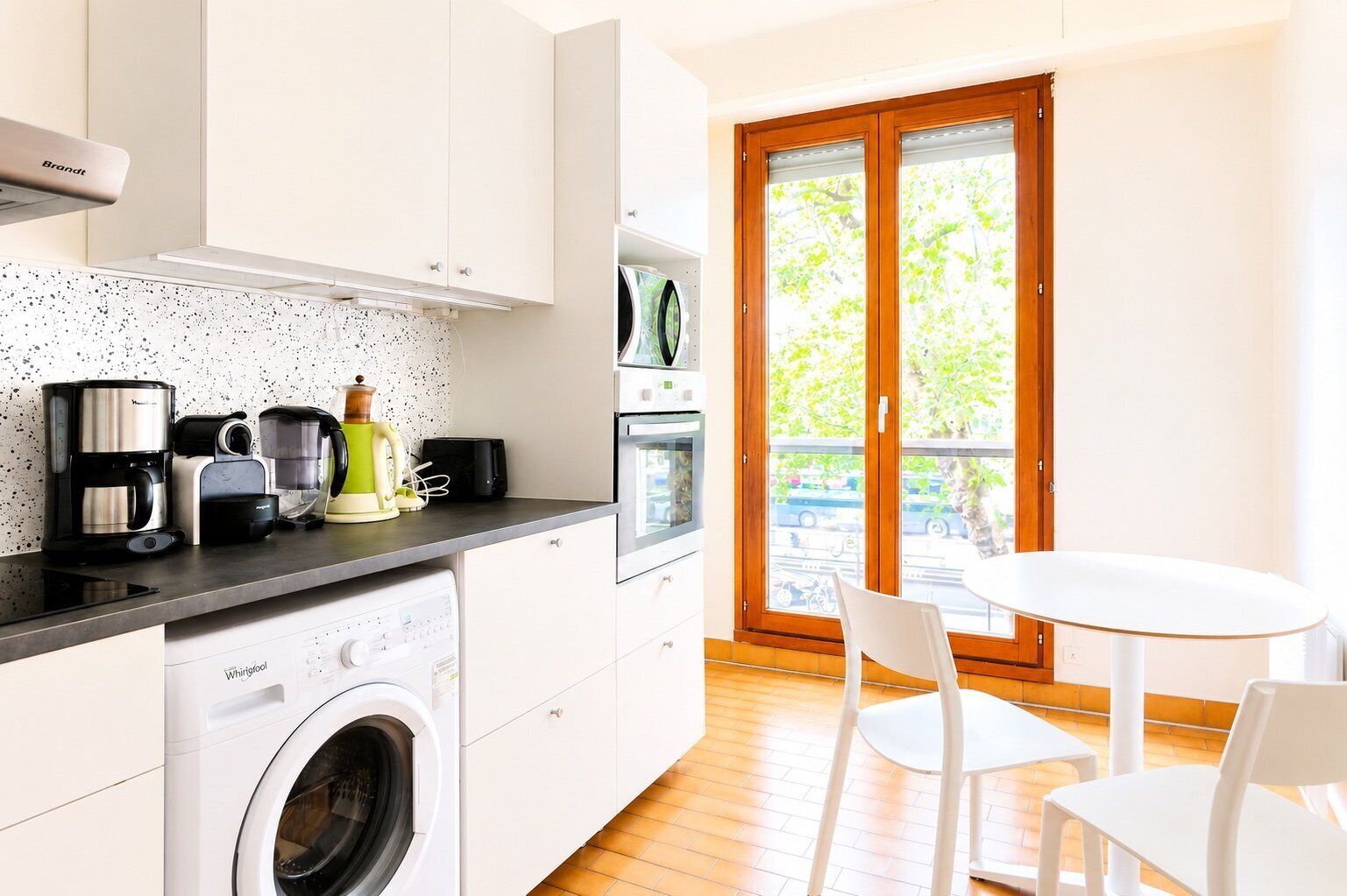 A compact kitchen with a washing machine, coffee maker, and toaster. There is a dining area with a table and chairs, and a sliding glass door leading to an outdoor area.
