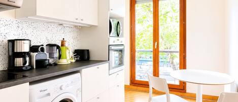 A compact kitchen with a washing machine, coffee maker, and toaster. There is a dining area with a table and chairs, and a sliding glass door leading to an outdoor area.