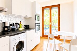 A compact kitchen with a washing machine, coffee maker, and toaster. There is a dining area with a table and chairs, and a sliding glass door leading to an outdoor area.