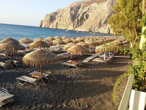 A beach with rows of sun loungers and thatched umbrellas, a rocky cliff in the background, and a clear view of the sea. - Ocean View (Santorini)