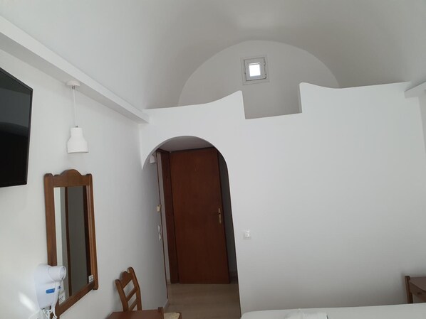 A bedroom with a bed, a desk with a chair, a mirror, a ceiling light, and a door leading to another room.