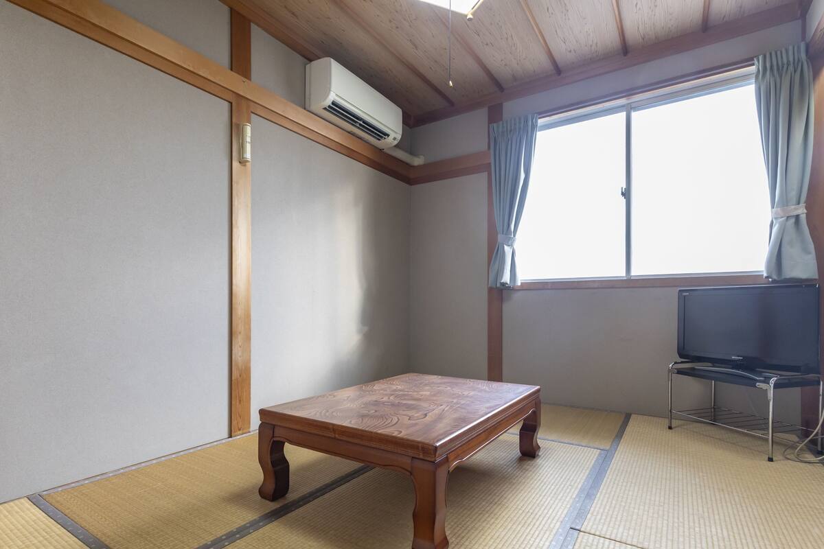 Una habitación con suelo de tatami, una mesa de madera, un televisor sobre un mueble bajo y una ventana con cortinas.
