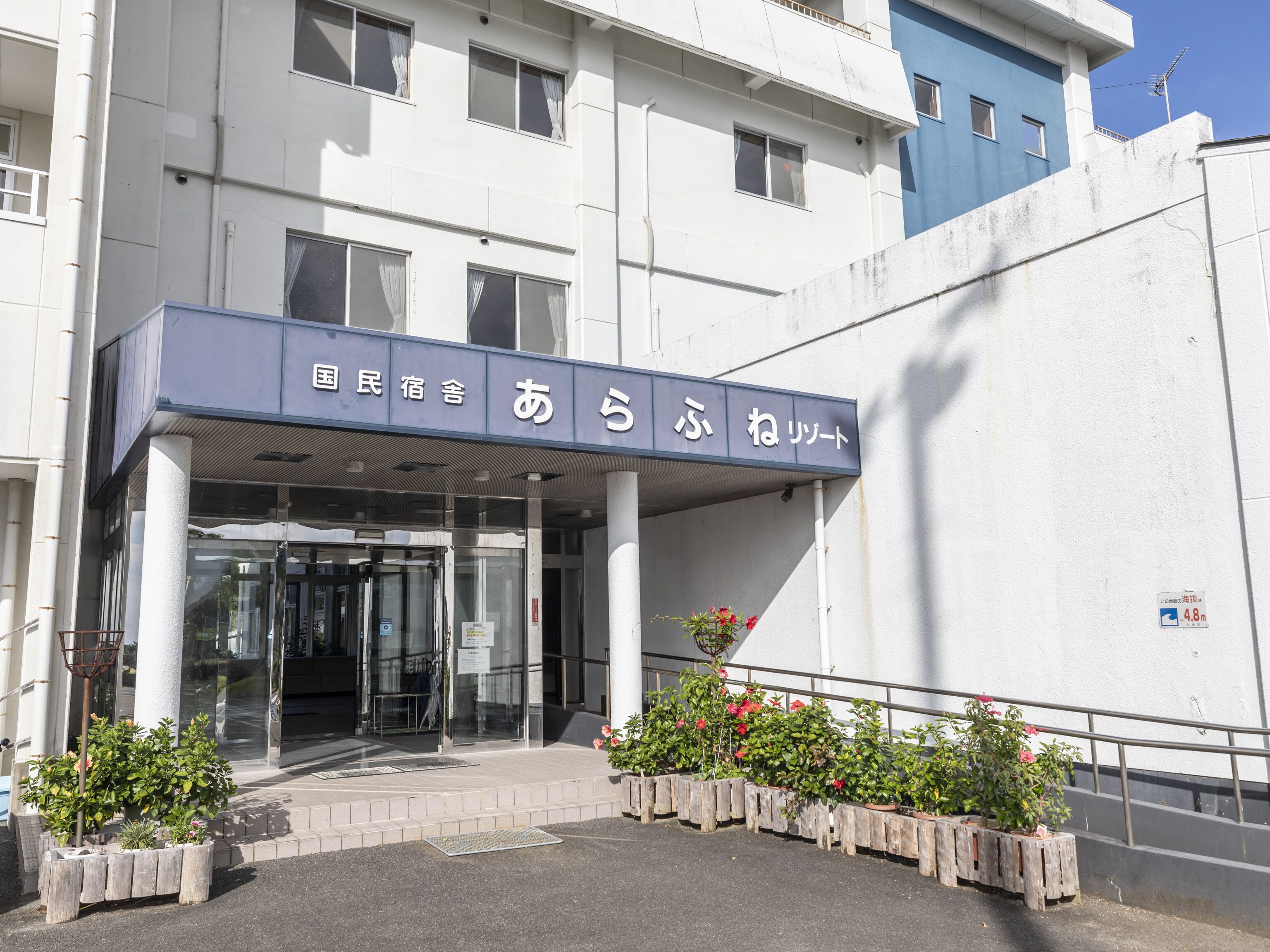 A building entrance with a covered entryway, a sign with Japanese characters, and potted plants.