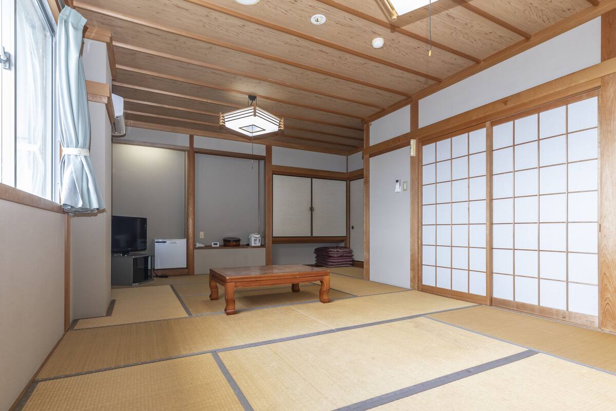 Una habitación japonesa tradicional con suelo de tatami, una mesa de madera, puertas correderas y un ventilador de techo.