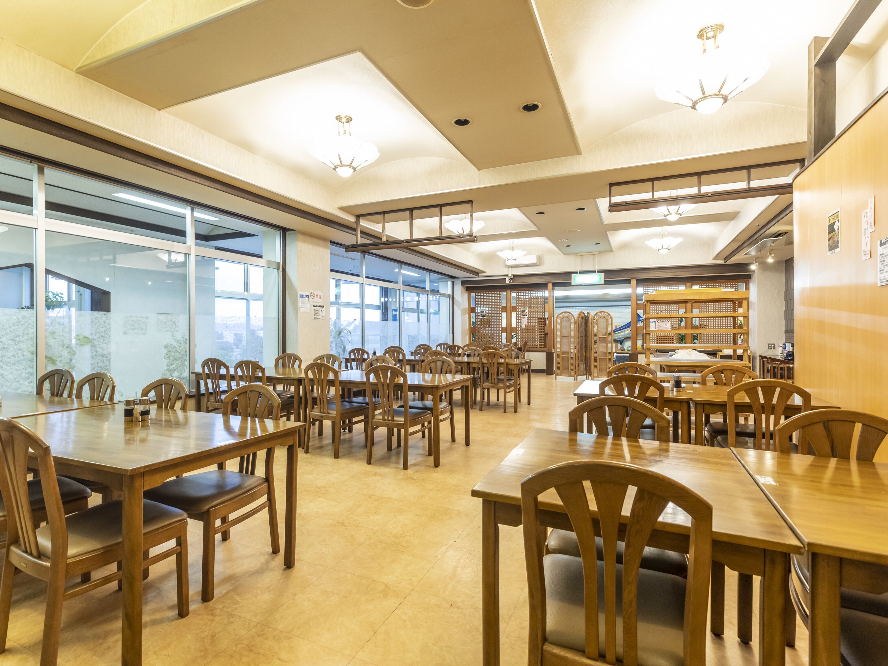 A spacious indoor area with multiple wooden tables and chairs, large windows allowing natural light, and a ceiling with recessed lighting.