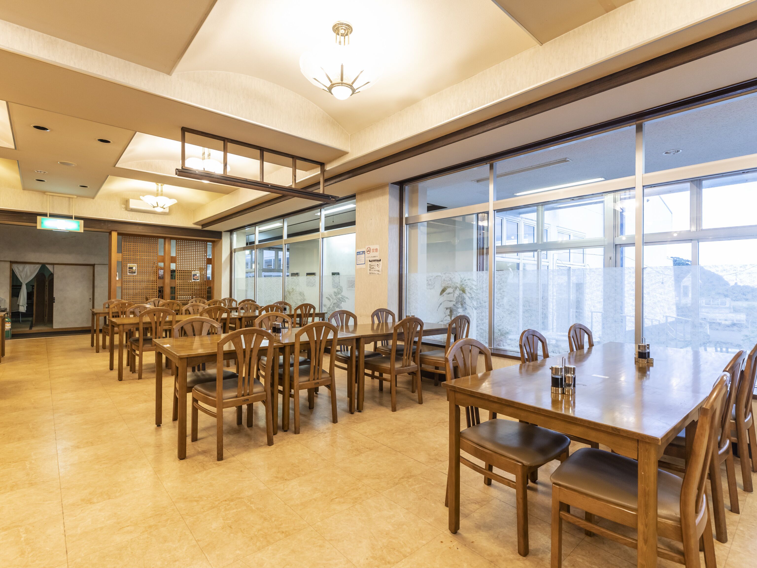 Un vaste espace dédié à la salle à manger, avec des tables et des chaises en bois, de grandes fenêtres offrant une vue sur l’extérieur et un plafond doté d’un éclairage encastré.