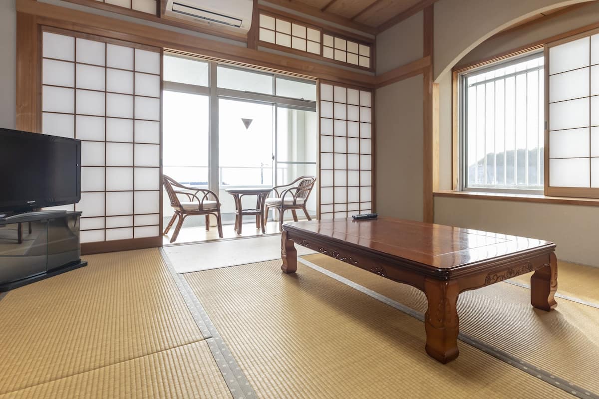 Una habitación japonesa tradicional con suelo de tatami, una mesa de madera baja, dos sillas, un televisor y ventanas amplias con biombos shoji.