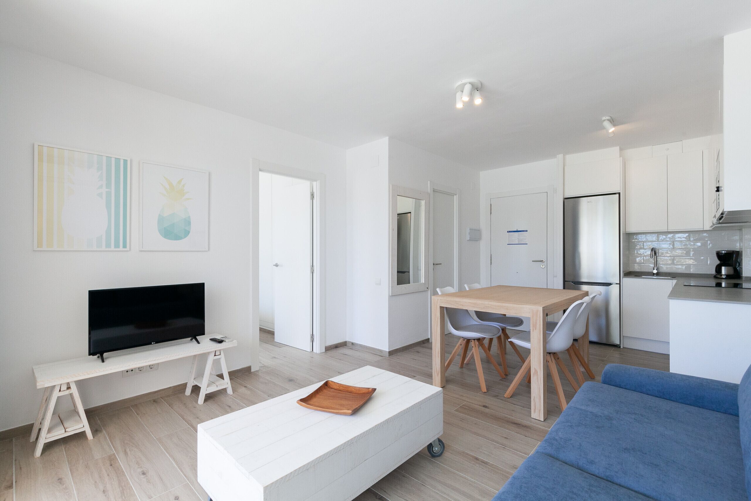 Una sala moderna con un televisor de pantalla plana, un sofá blanco, una mesa de comedor de madera con sillas, y una cocina con alacenas blancas y un refrigerador de acero inoxidable.