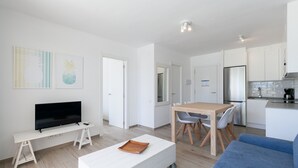 A modern living room with a flat-screen TV, a white sofa, a wooden dining table with chairs, and a kitchen area with white cabinets and a stainless steel refrigerator. - Beachfront Apartment · AirCon · WiFi · Pool UHC NOVELTY 010 (Salou)