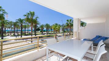 Un balcón con una mesa y sillas blancas, con vista a una playa con palmeras y cielo despejado.
