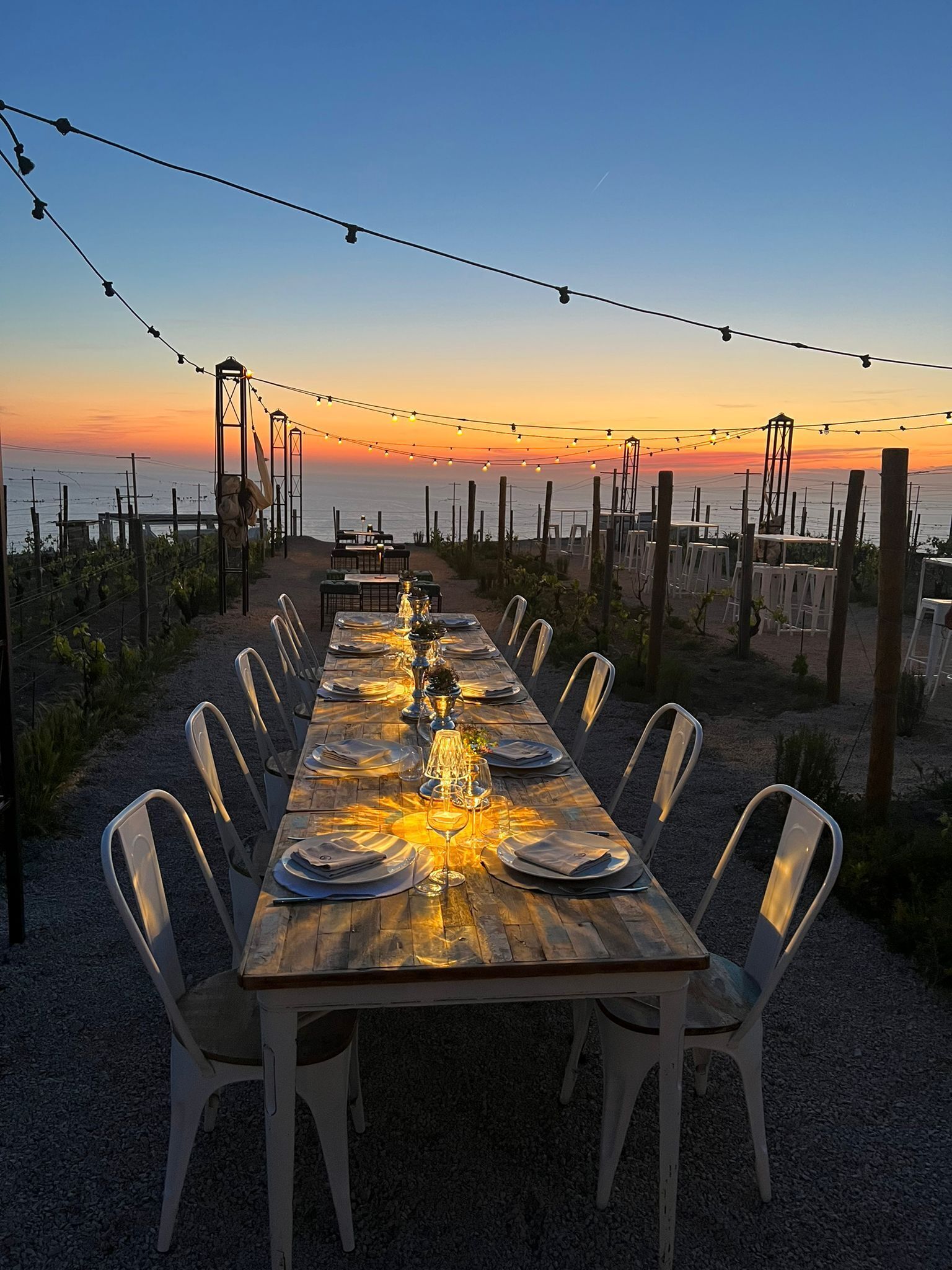 Una larga mesa de comedor al aire libre con sillas, luces colgantes y vista al océano al atardecer.