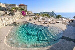 Zona de piscina con agua cristalina, rodeada de sillas de descanso y sombrillas, con vistas a un paisaje costero.