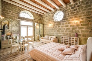 Un dormitorio con una cama grande, una mesa de comedor de vidrio y un sofá. Hay dos ventanas arqueadas y una pared de piedra.