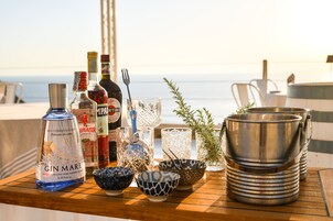 Una mesa con varias botellas, vasos y recipientes, probablemente para un cóctel o aperitivo, frente a un balcón con vista al mar.