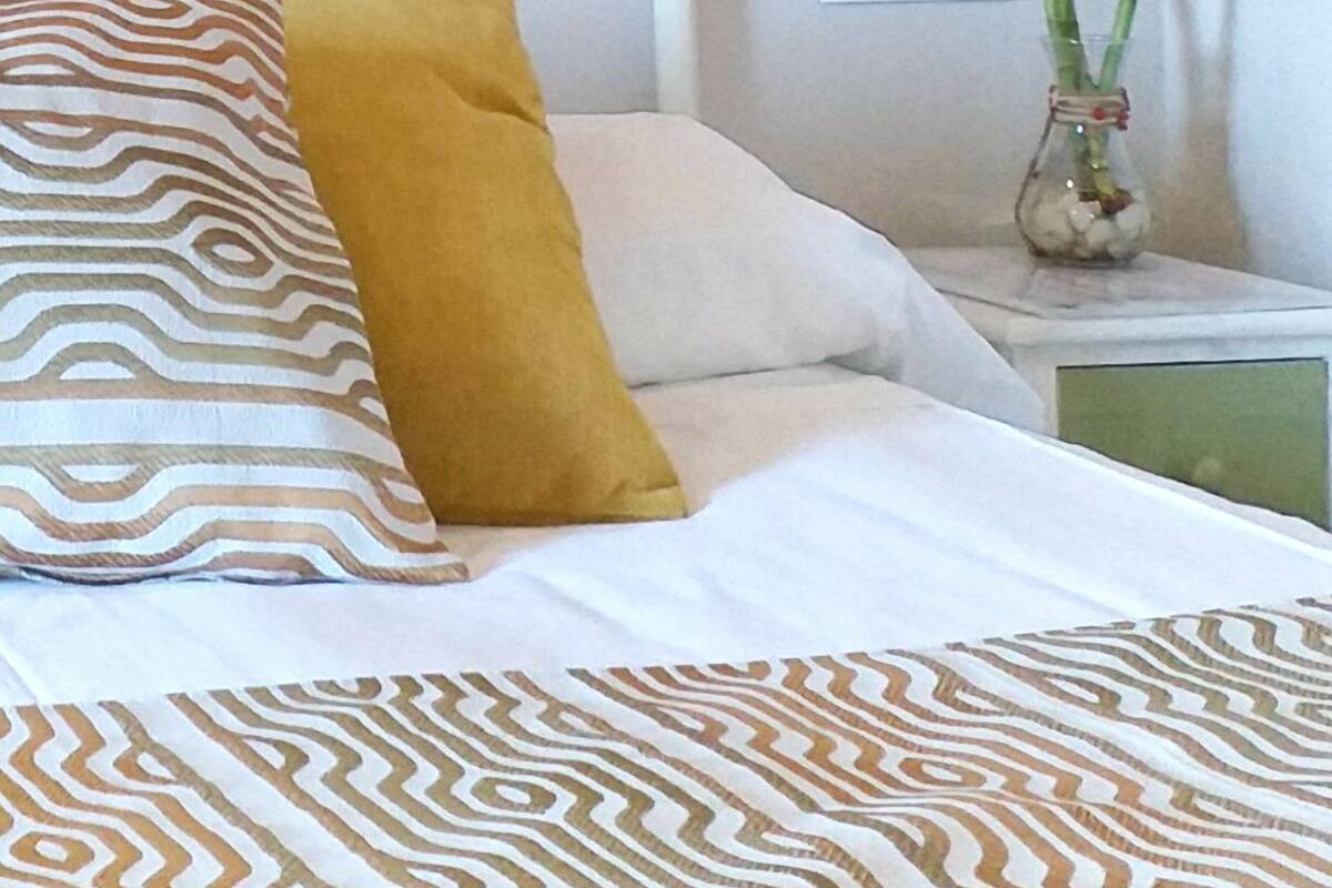 A neatly made bed with patterned bedding, a white headboard, and a bedside table with a vase of green plants.