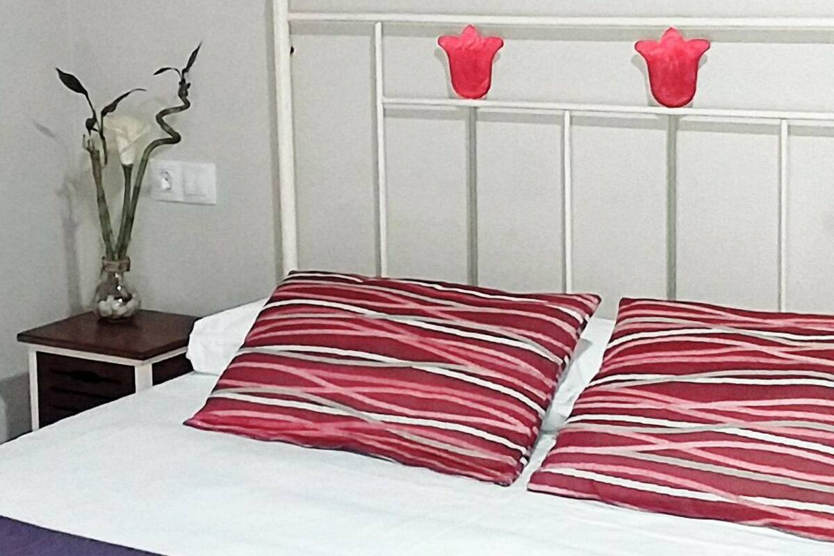 A bed with white bedding and red striped pillows, a small nightstand with a vase of flowers, and a white headboard with two red flower decorations.