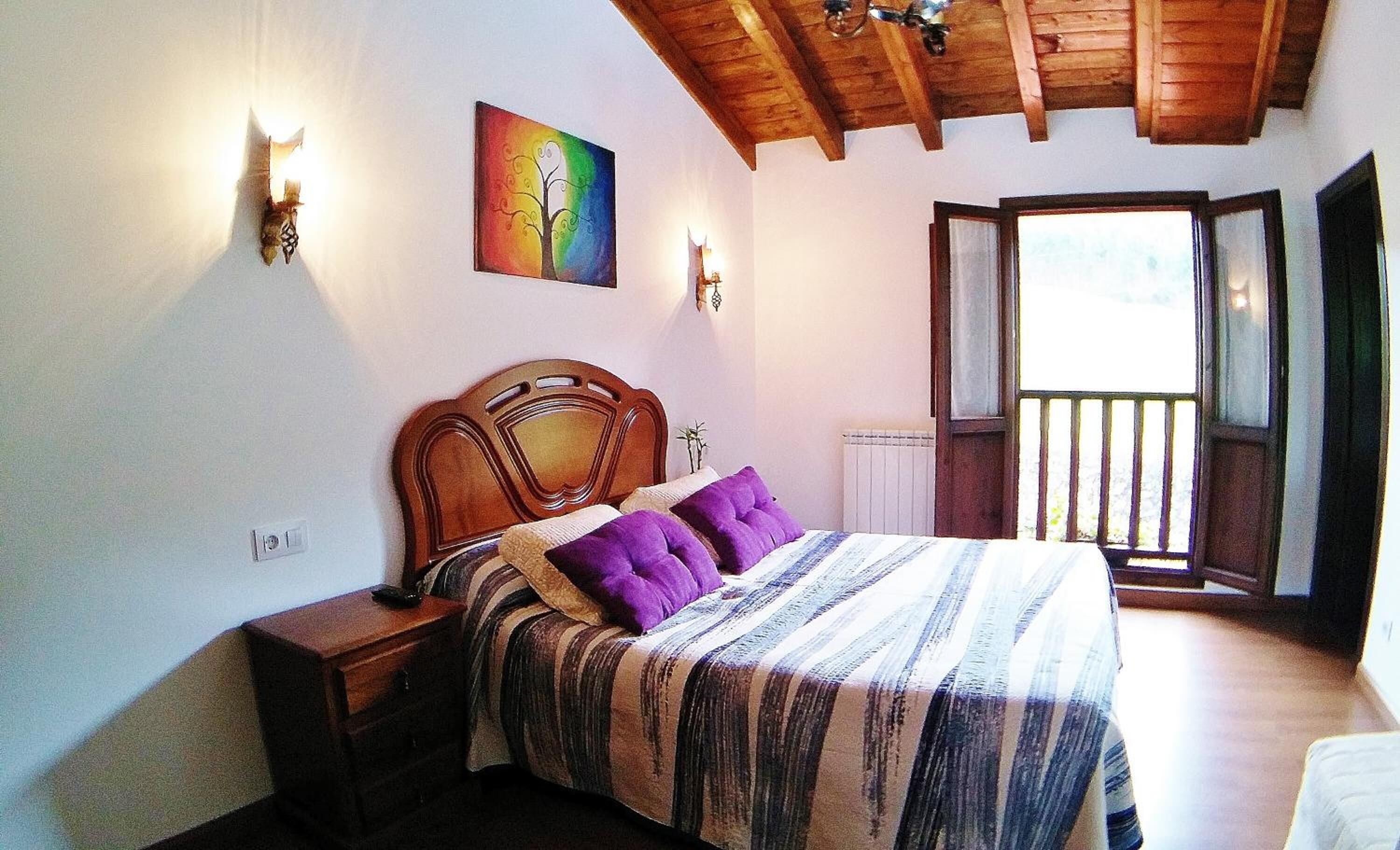 Un dormitorio con techo de madera, cama con ropa de cama a rayas y almohadas moradas, una mesita de noche, una lámpara de pared y una ventana con vistas.