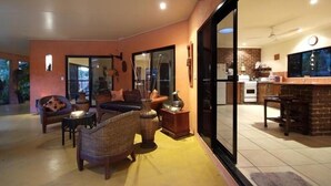 Un salon chaleureux avec un canapé, des fauteuils et une table basse. Une porte-fenêtre coulissante donne sur une cuisine dotée d’un mur de fond en briques et d’appareils électroménagers modernes.