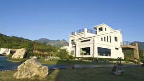 Une maison moderne à deux étages, dotée d’un balcon, entourée d’un jardin bien entretenu et de gros rochers.