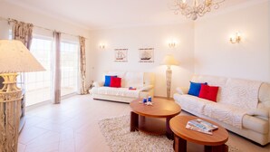 A spacious living room with two white sofas, a chandelier, and a large window with curtains.