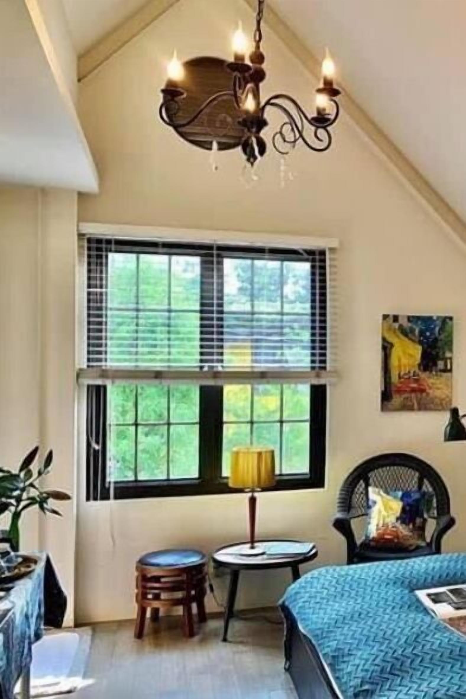 Une pièce avec un plafond en pente, un lustre, une fenêtre avec des stores, un tableau, un lit recouvert d’une couverture bleue, une chaise et une petite table.