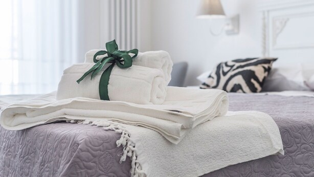 Una cama bien hecha con toallas blancas dobladas y un lazo verde, una colcha morada estampada y una almohada con estampado de cebra en una habitación luminosa con cabecera blanca y una lámpara de pared.