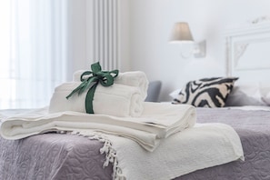 Una cama bien hecha con toallas blancas dobladas y un lazo verde, una colcha morada estampada y una almohada con estampado de cebra en una habitación luminosa con cabecera blanca y una lámpara de pared.