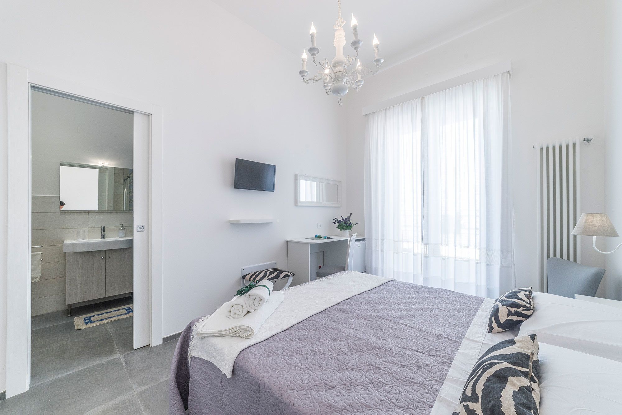 Une chambre d’hôtel moderne, équipée d’un lit, d’un bureau, d’une télévision et d’une salle de bain visible à travers une porte ouverte.
